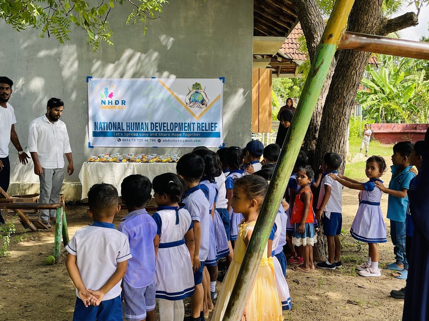 Schoolchildren lined up beside an NHDR banner and a table of fruit and yoghurt