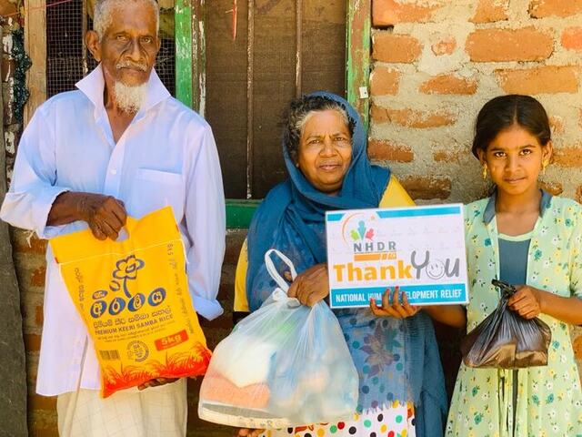 A family sheltering under a palm-thatch roof receiving supplies from NHDR