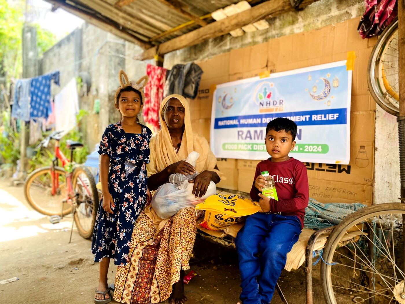 A mother and two children with an NHDR Ramadan food pack outside their home